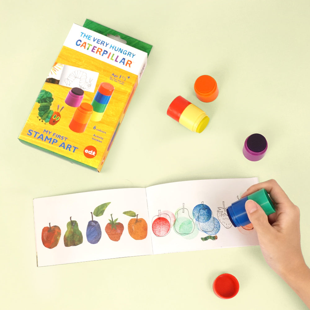 The Very Hungry Caterpillar 'My First Stamp Art' kit with colorful stampers and a child using a blue stamp to create fruit art in a workbook.