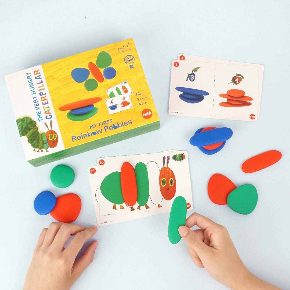 The Very Hungry Caterpillar 'My First Rainbow Pebbles' educational toy set with colorful pebbles, activity cards, and a child's hands arranging pieces to match a caterpillar pattern.